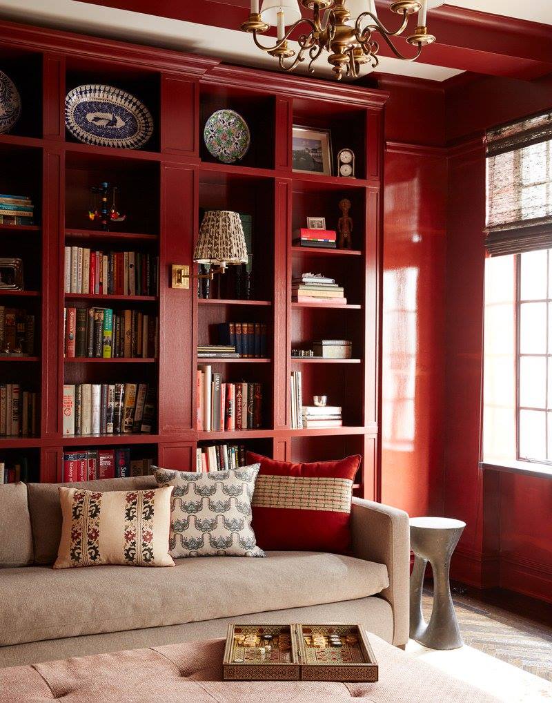 Cozy library with rich red built-in shelves, beige sofa adorned with patterned pillows, brass chandelier, and natural light from large windows.
